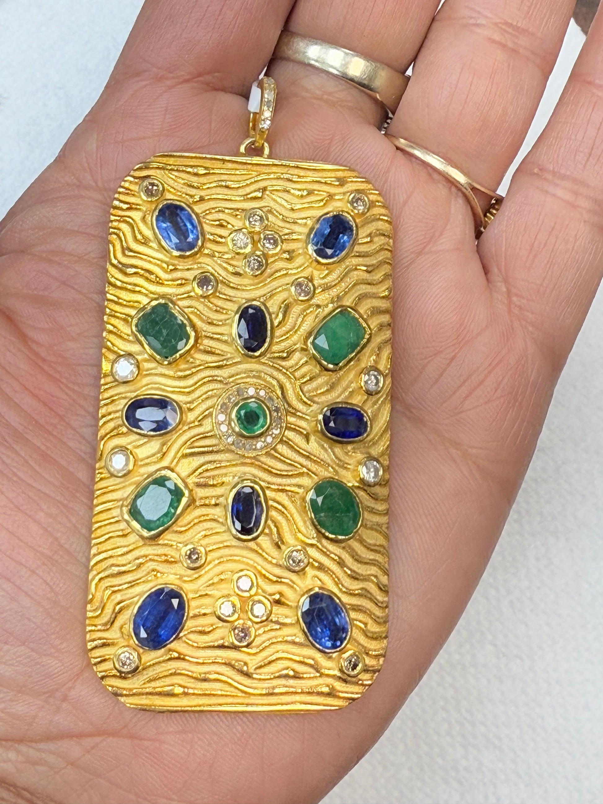 A gold-colored pendant with diamond and gemstone pave charms, held in a person's hand.