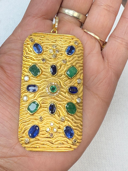 A gold-colored pendant with diamond and gemstone pave charms, held in a person's hand.